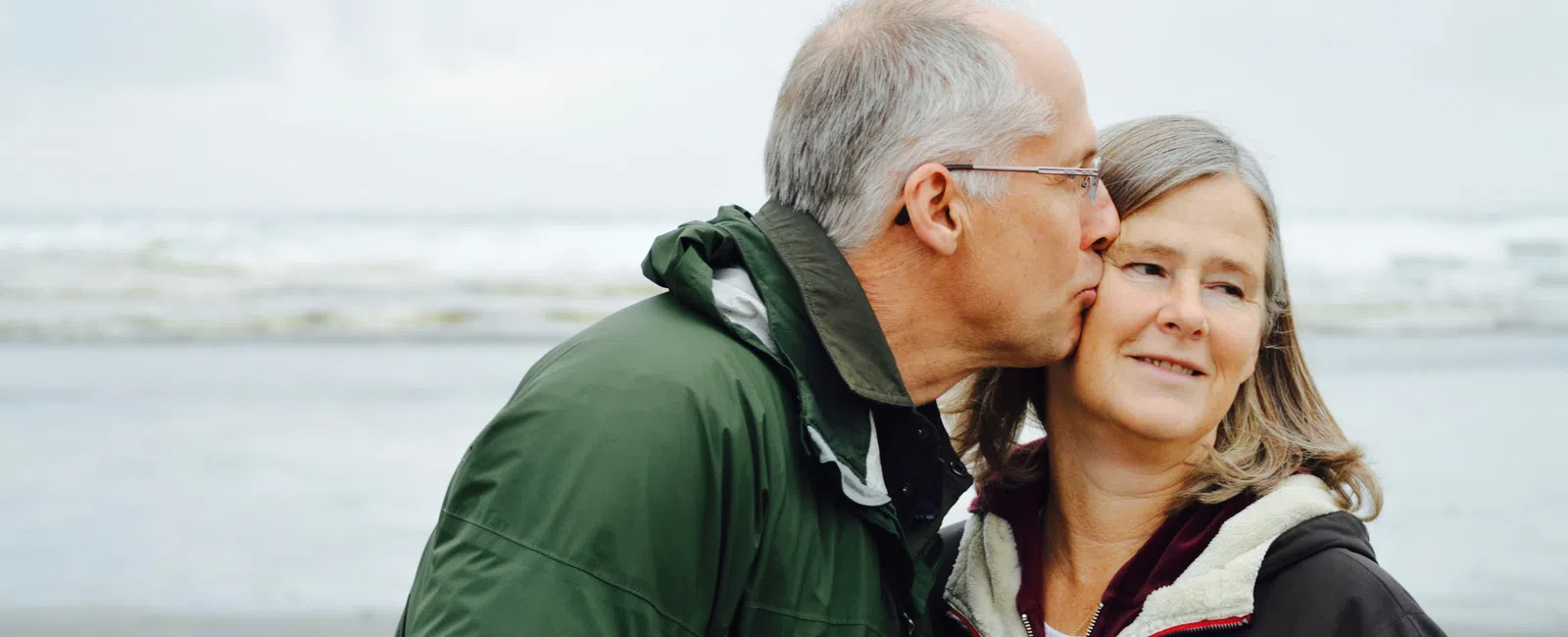 Mature man kisses his smiling partner on the cheek by the beach on an overcast day.