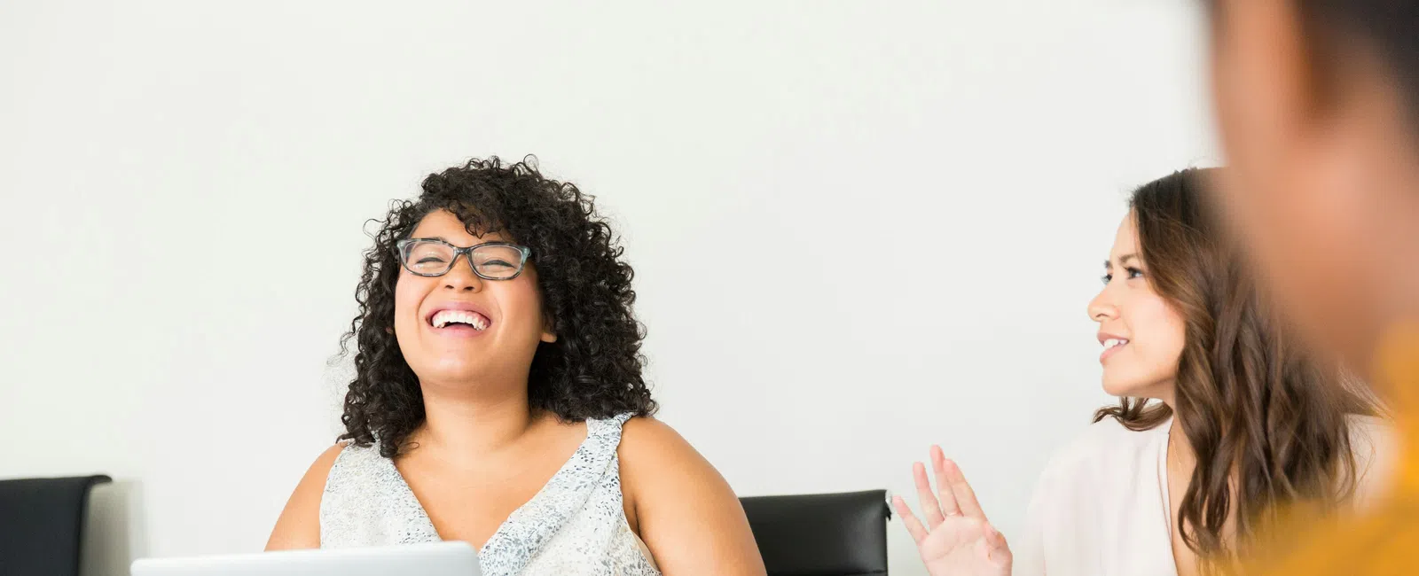 Smiling woman with curly hair using a laptop during a lively office meeting with colleagues.