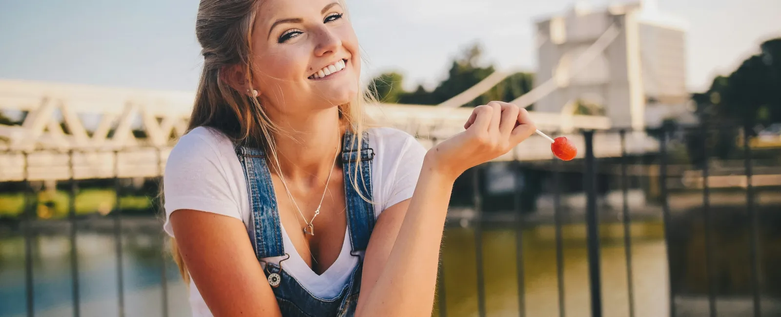 Smiling young woman in denim overalls holding a red lollipop outdoors by a railing near water at sunset.