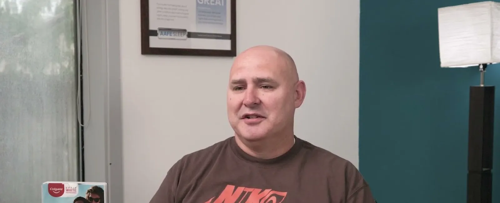 Man in brown Nike Air t-shirt sitting in a room with dental product boxes and a framed certificate on the wall.