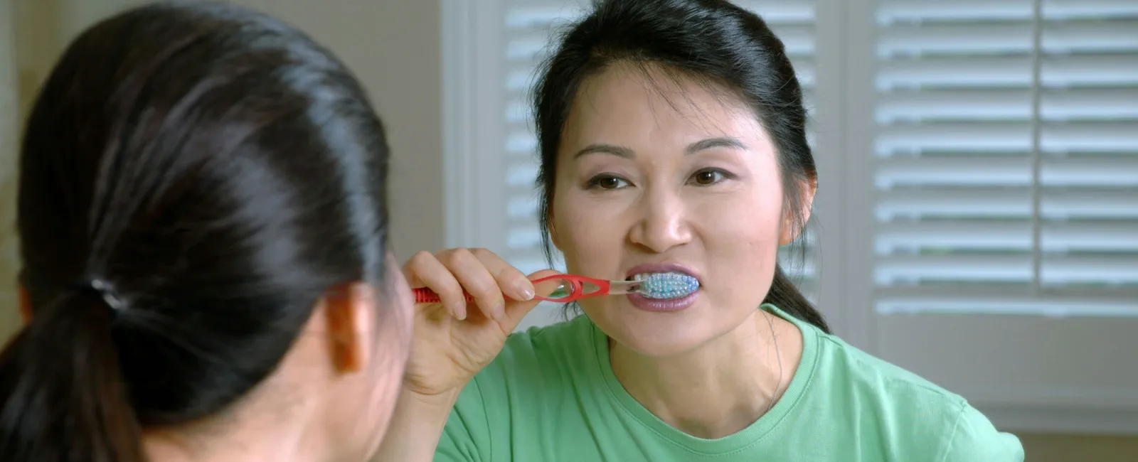 Woman in green shirt brushing teeth while looking in the bathroom mirror with natural light.