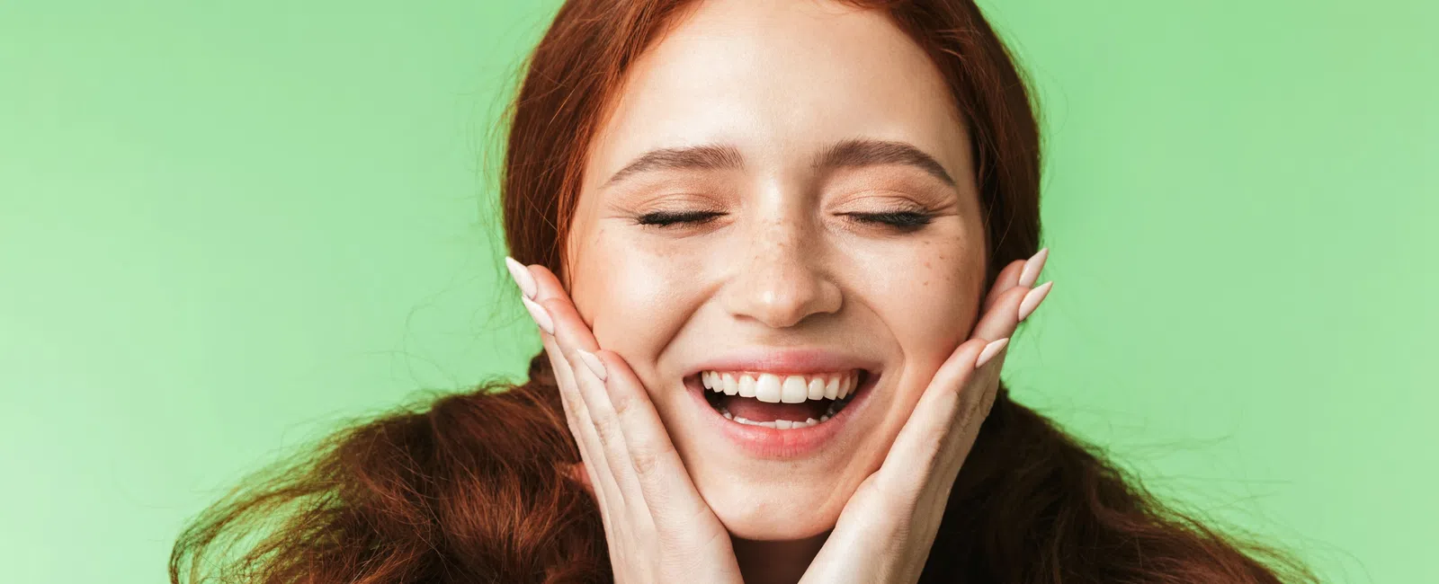 Smiling red-haired woman with closed eyes and hands on cheeks against a green background