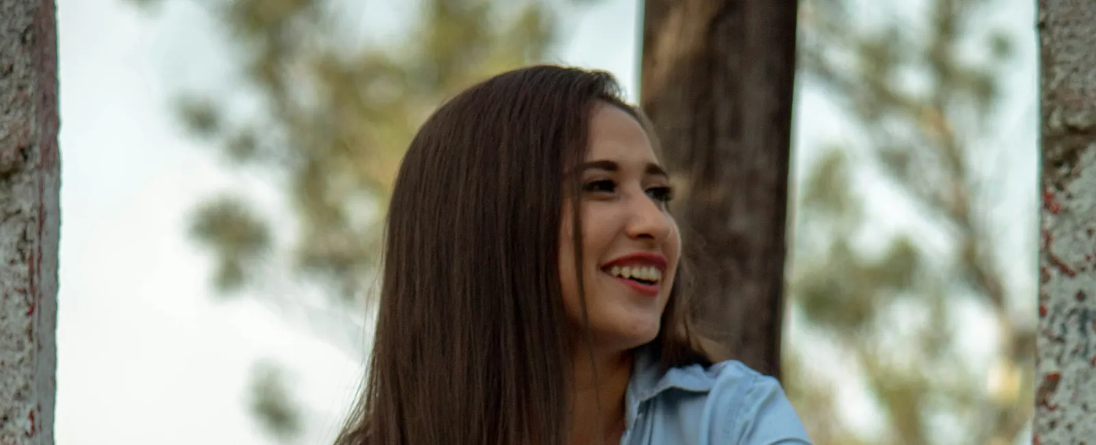 Smiling woman in ripped jeans and denim shirt sitting on weathered stone steps outdoors with greenery.