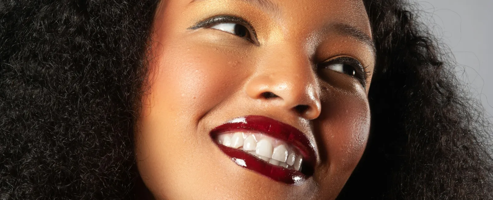 Smiling woman with natural curly hair wearing glossy dark red lipstick and gold eyeshadow on a gray background