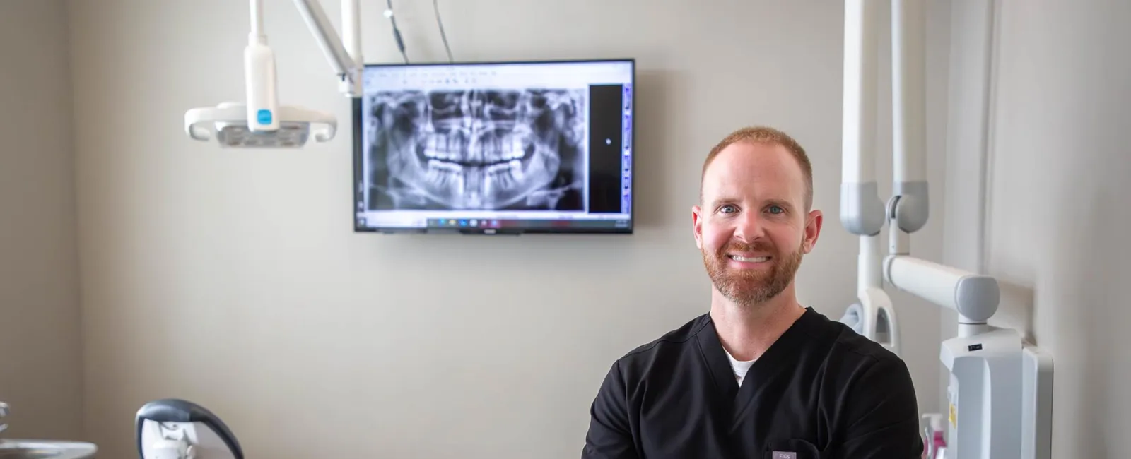 Smiling male dentist in black scrubs stands confidently in dental office with panoramic dental X-ray on screen