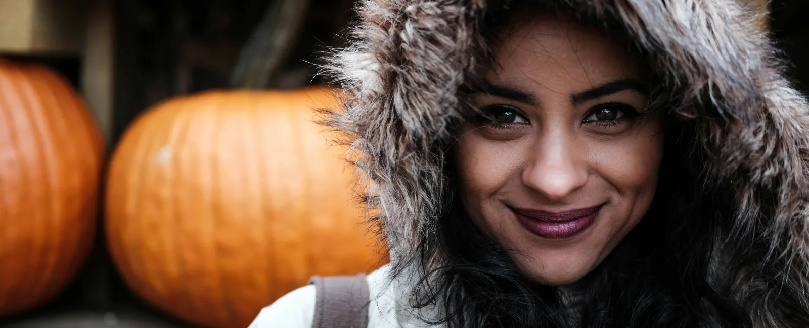 Woman in a fur-lined hooded coat smiles with pumpkins displayed on wooden shelves behind her.