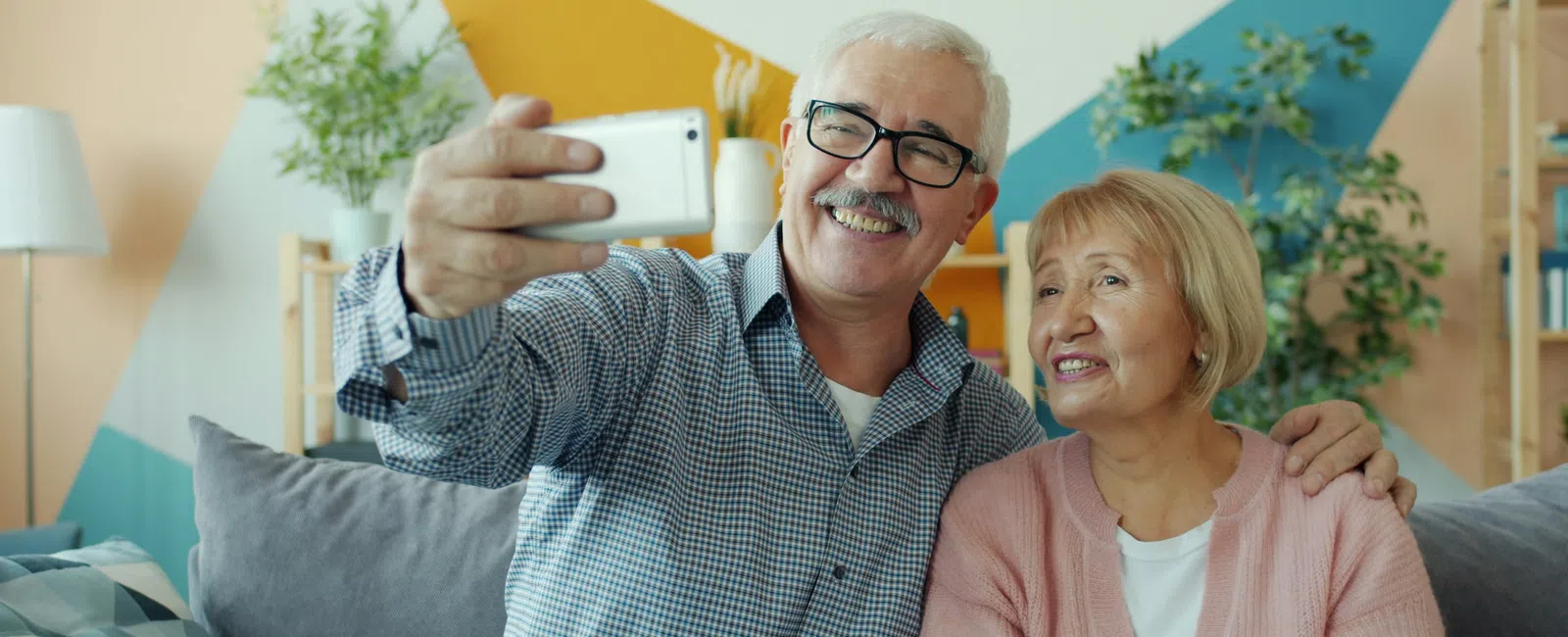 Senior couple sitting on couch taking a happy selfie in a cozy living room with colorful wall art.