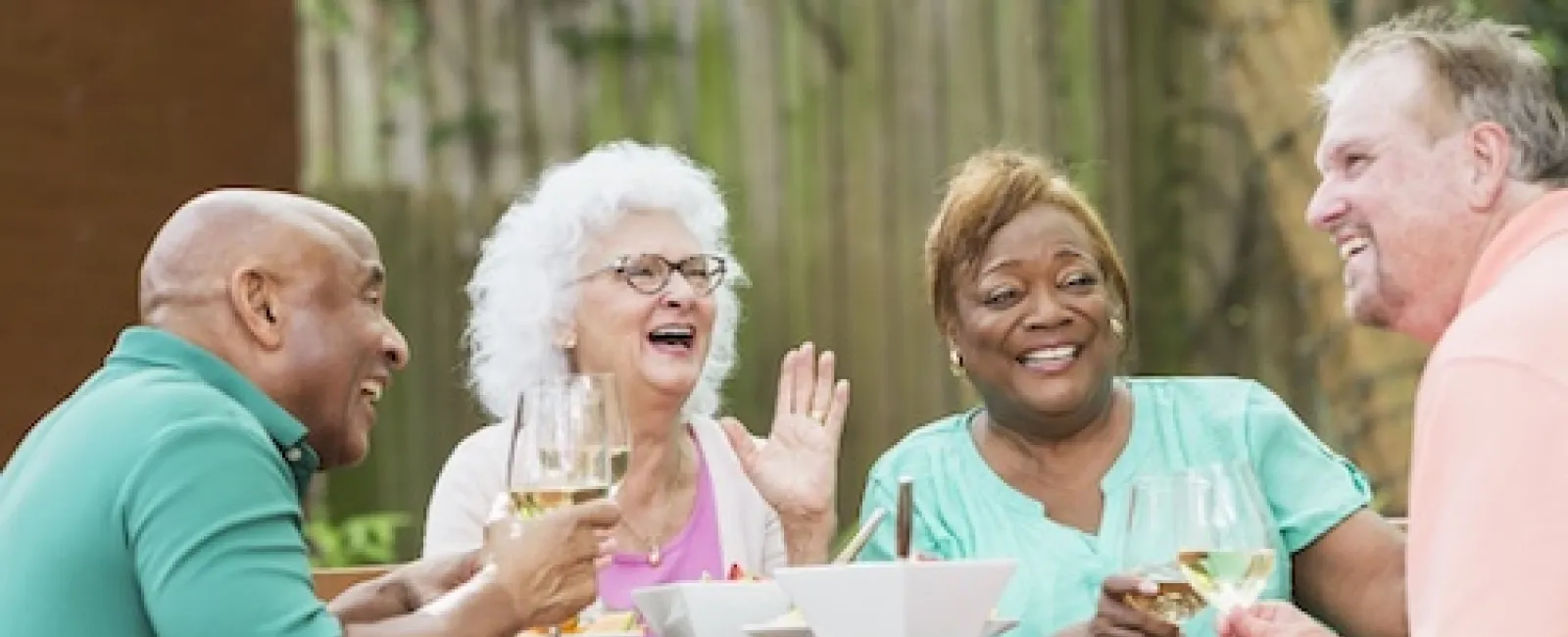 Four diverse senior friends enjoying a cheerful outdoor meal with white wine and conversation around a wooden table.