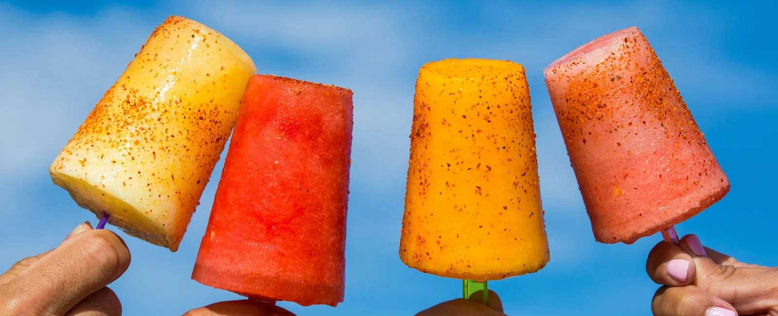Four colorful fruit popsicles sprinkled with chili powder held against a bright blue sky background.