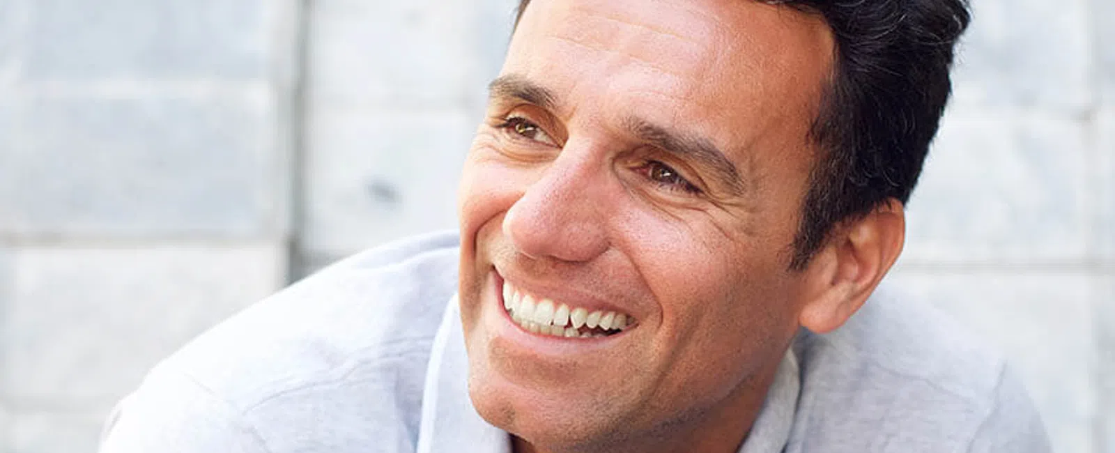 Smiling man with curly hair in a gray polo shirt against a textured wall background.