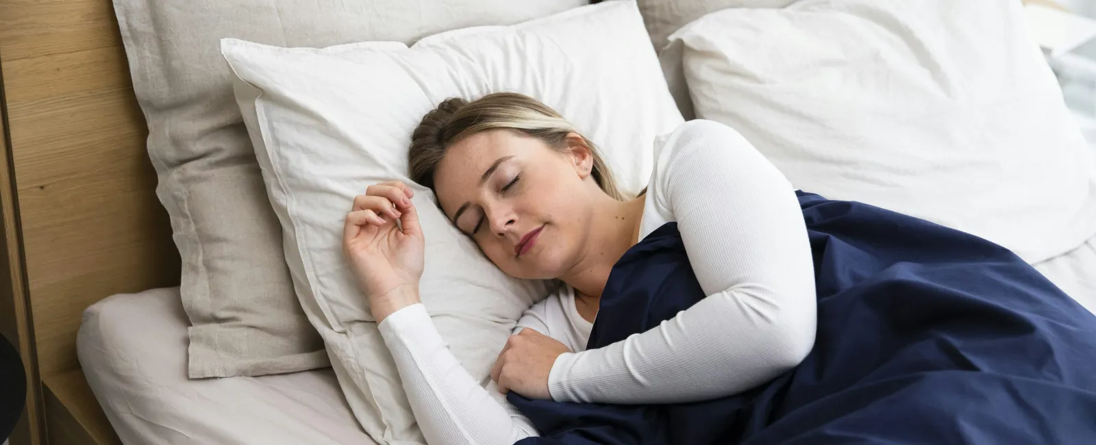 Woman peacefully sleeping on white pillows in a wooden bed with beige sheets and a navy blue blanket.
