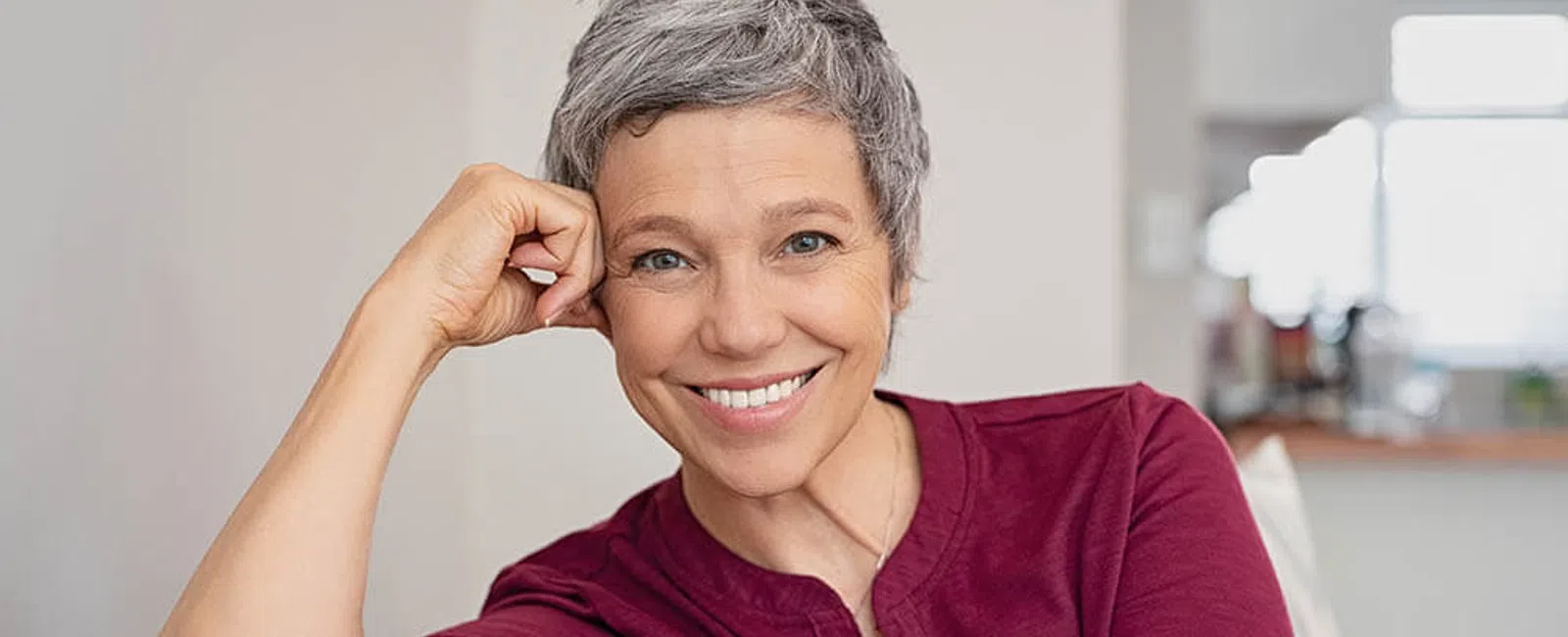 Smiling mature woman with short gray hair relaxing on a couch wearing a burgundy top and silver watch.
