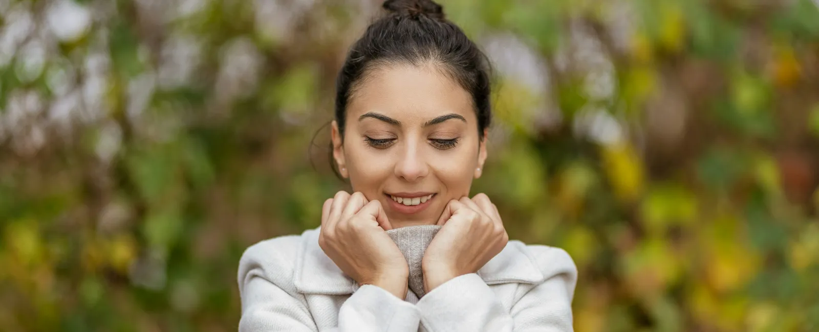 Young woman in a white coat smiling and holding collar close outdoors with blurred green and brown background.