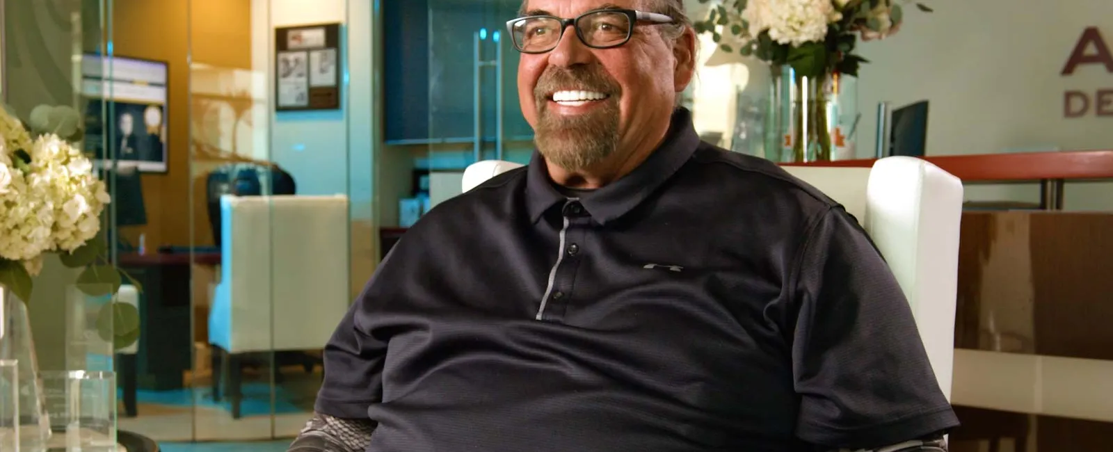 Smiling middle-aged man with glasses sitting in a modern office with flowers and glass walls.