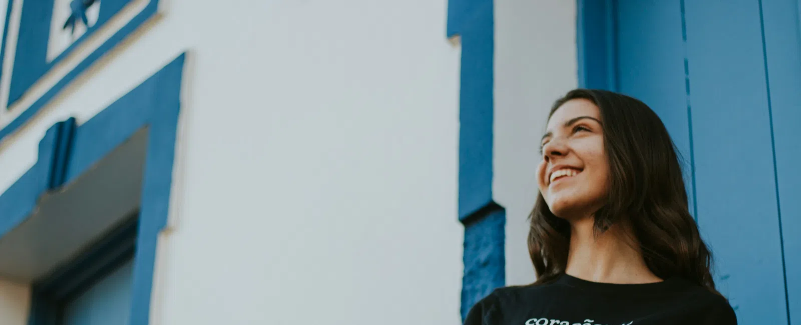 Smiling woman in casual black t-shirt stands near white building with blue door and trim outdoors.