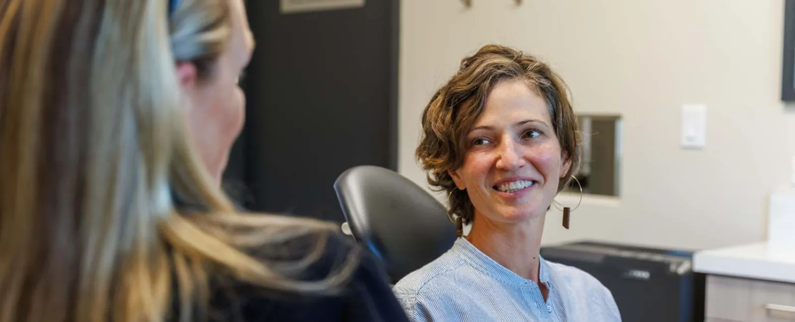 Two women in conversation in a modern office setting, one smiling and seated in a chair facing the other.