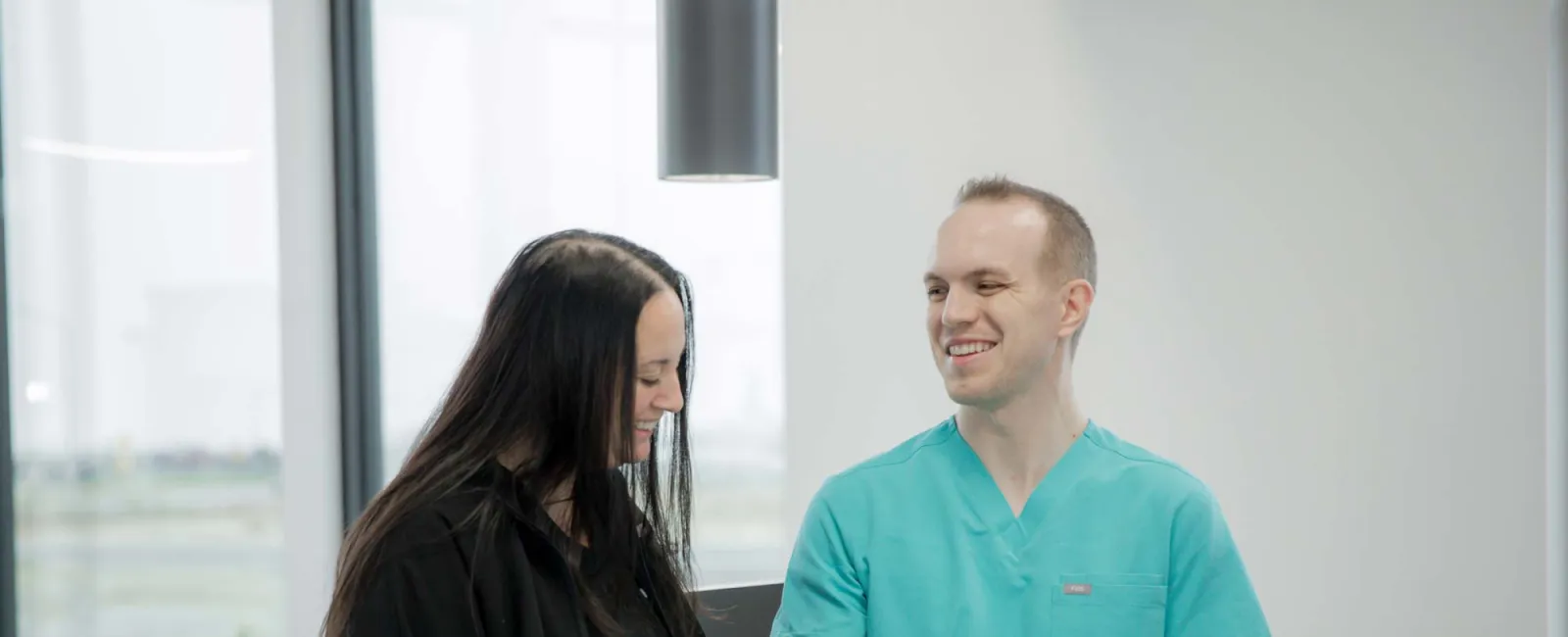 Healthcare professional in teal scrubs discussing information with woman in black in a bright modern office.