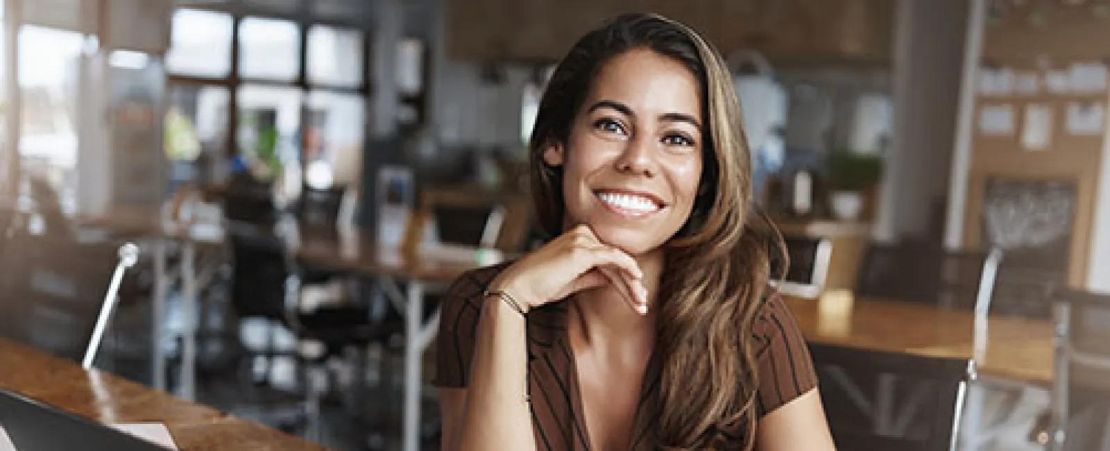 Smiling woman sitting at wooden desk with laptop, coffee cup, and papers in modern office space.