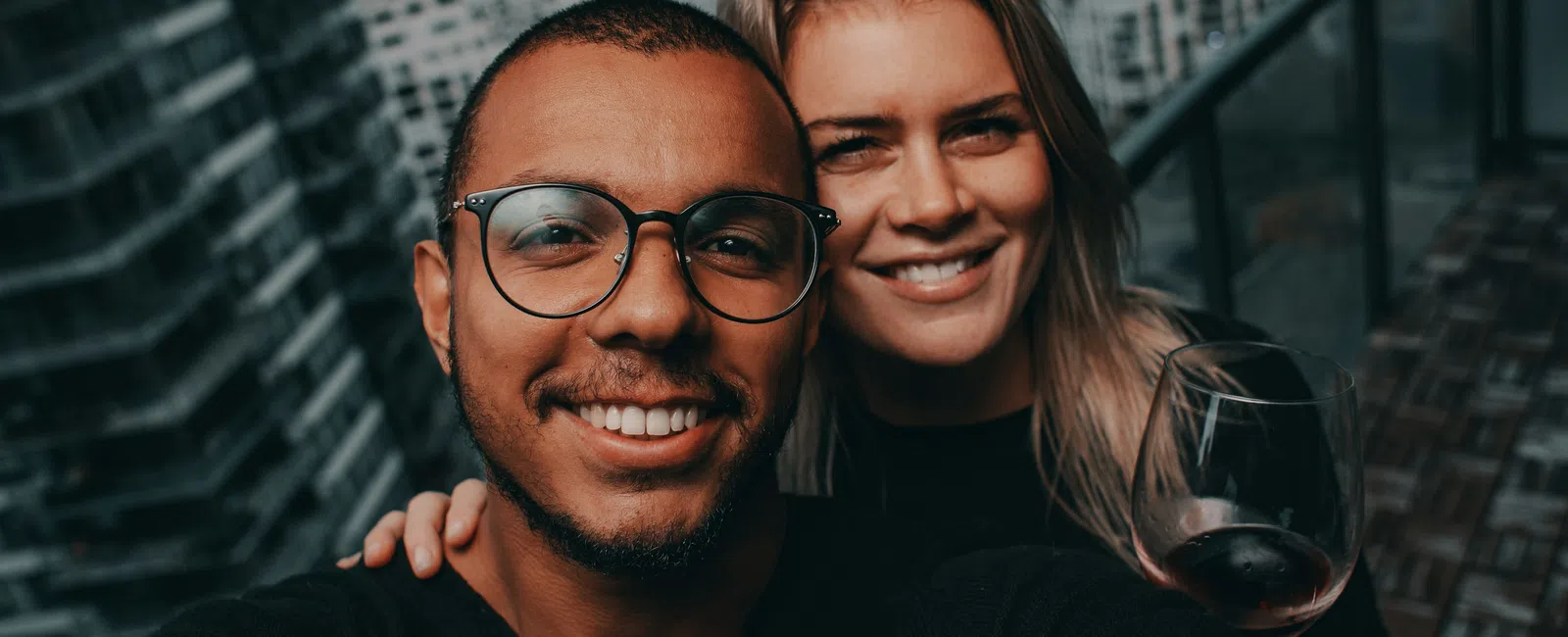 Smiling couple taking a selfie on a balcony with city buildings and ocean in the background, woman holding a glass of wine.
