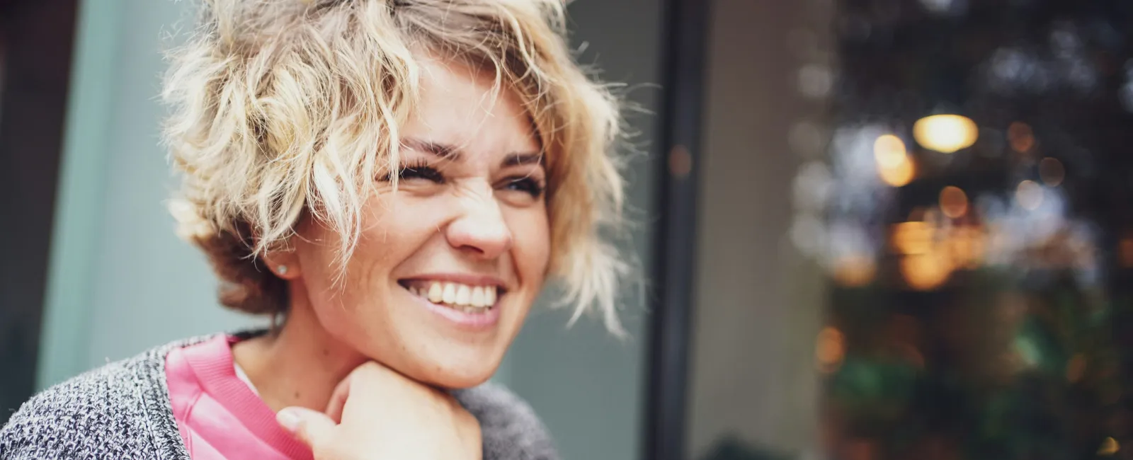 Smiling woman with short blonde hair wearing a pink sweatshirt and gray cardigan sitting outdoors near window.