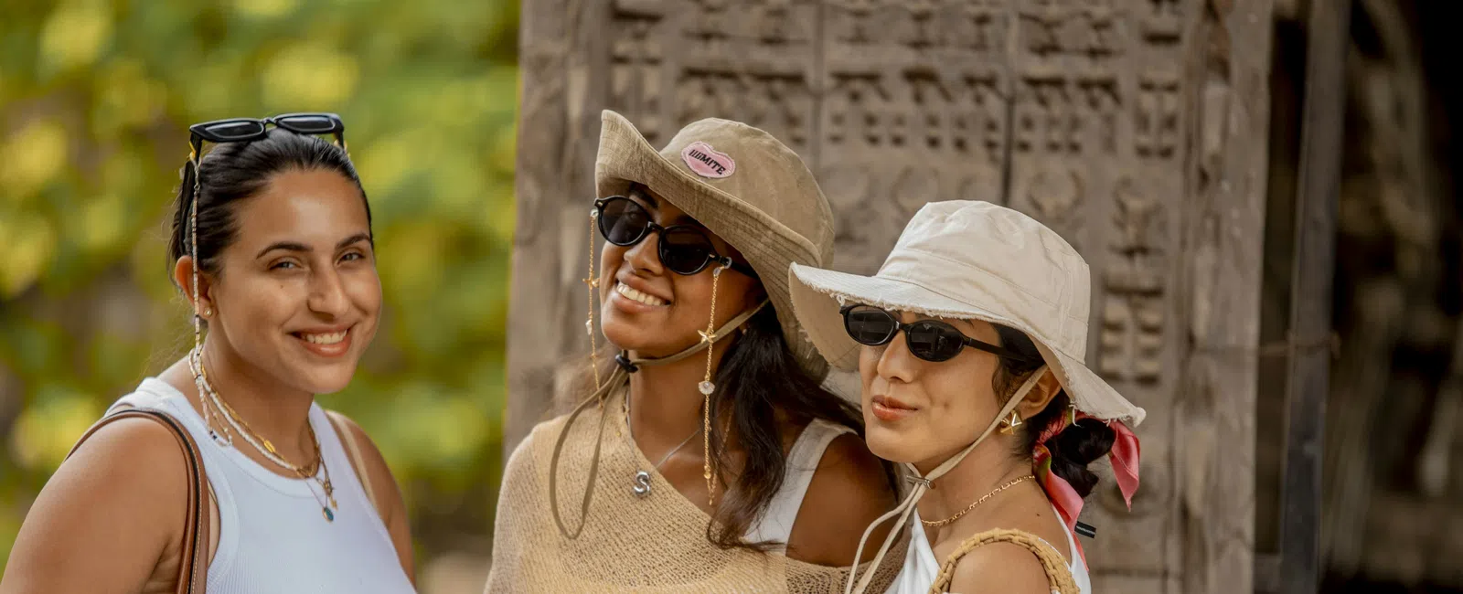 Three women wearing sunglasses and summer hats smiling outdoors with a carved stone background and greenery.