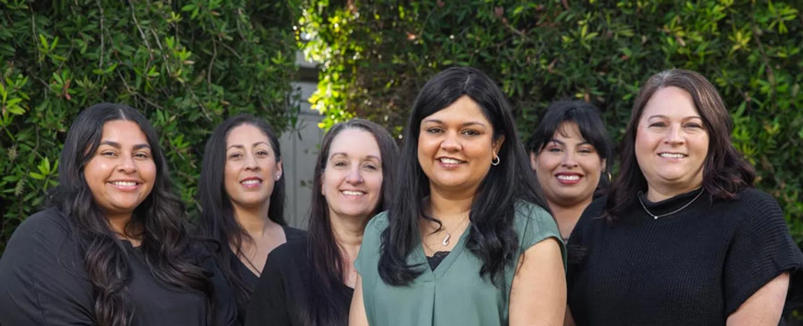 Group portrait of six diverse women standing outdoors smiling with greenery background in casual smart attire