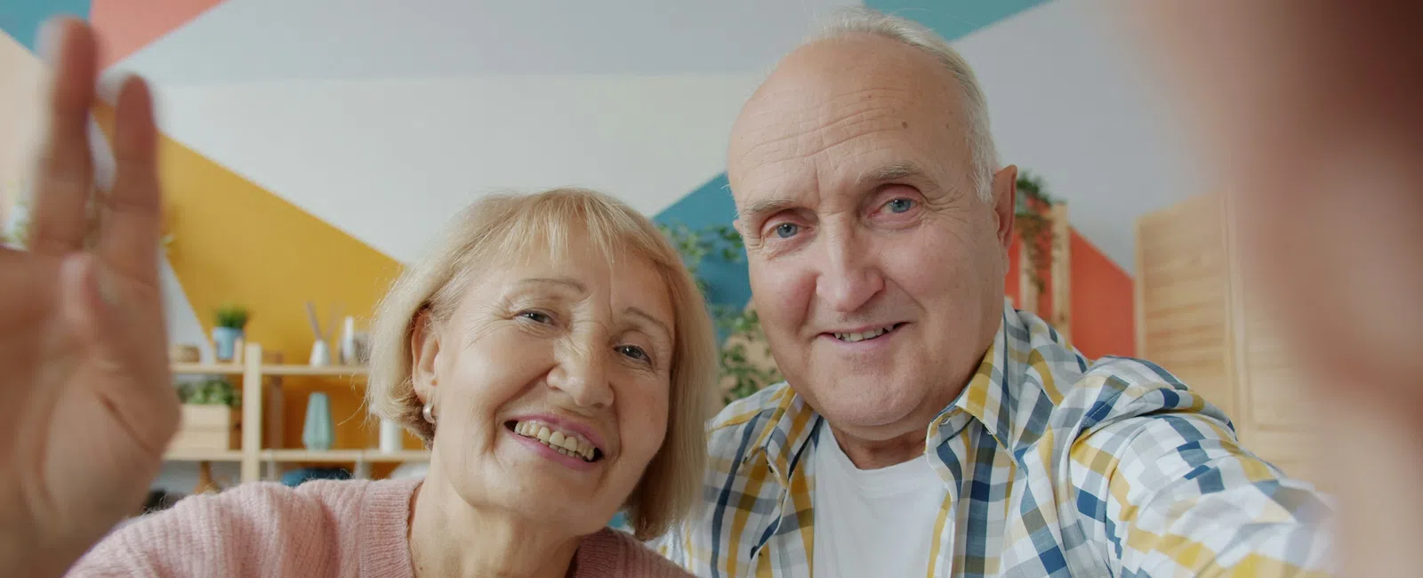 Smiling elderly couple taking a selfie together in a cozy, colorful living room setting.