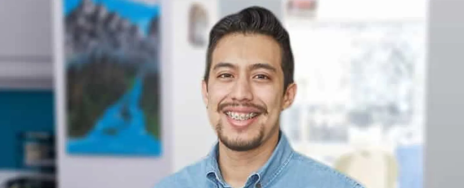 Smiling man with braces wearing a light blue denim shirt standing indoors with blurred background.