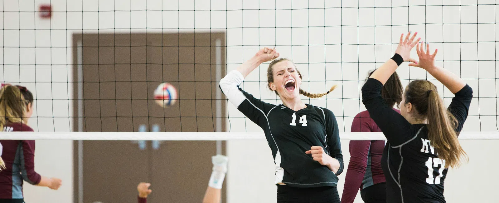 Female volleyball players in black and maroon uniforms competing intensely during a match indoors