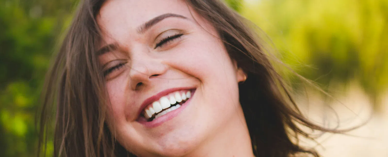 Happy young woman with brown hair smiling outdoors wearing a white lace top with greenery background