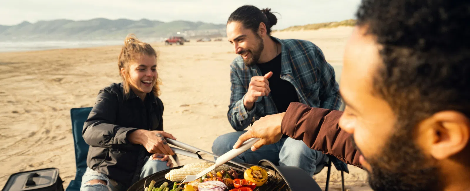 Three friends grilling mixed vegetables including peppers, corn, and asparagus on a portable grill at the beach.
