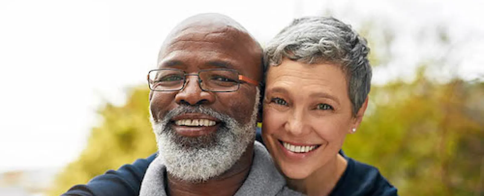 Smiling middle-aged couple outdoors, woman embraces man with grey beard and glasses on a bright day