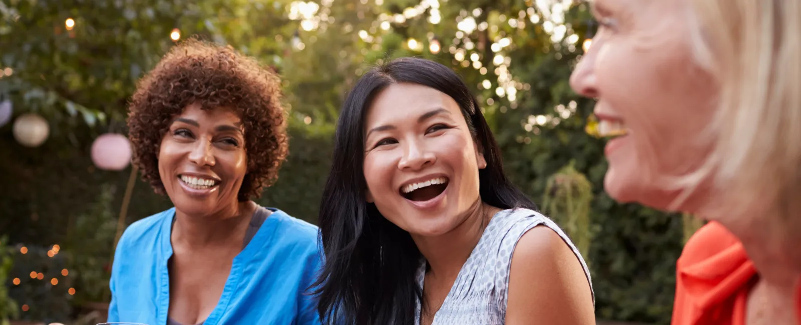 Three diverse women enjoying an outdoor meal together, laughing and sharing a joyful moment in a garden setting.