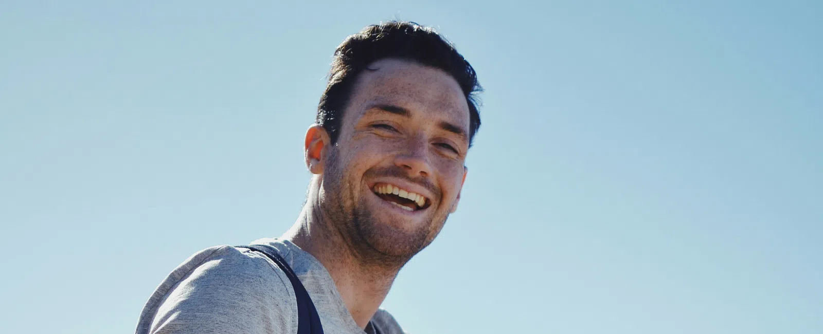 Smiling man in gray t-shirt and black shorts sitting on a rock outdoors wearing a backpack and watch on a sunny day