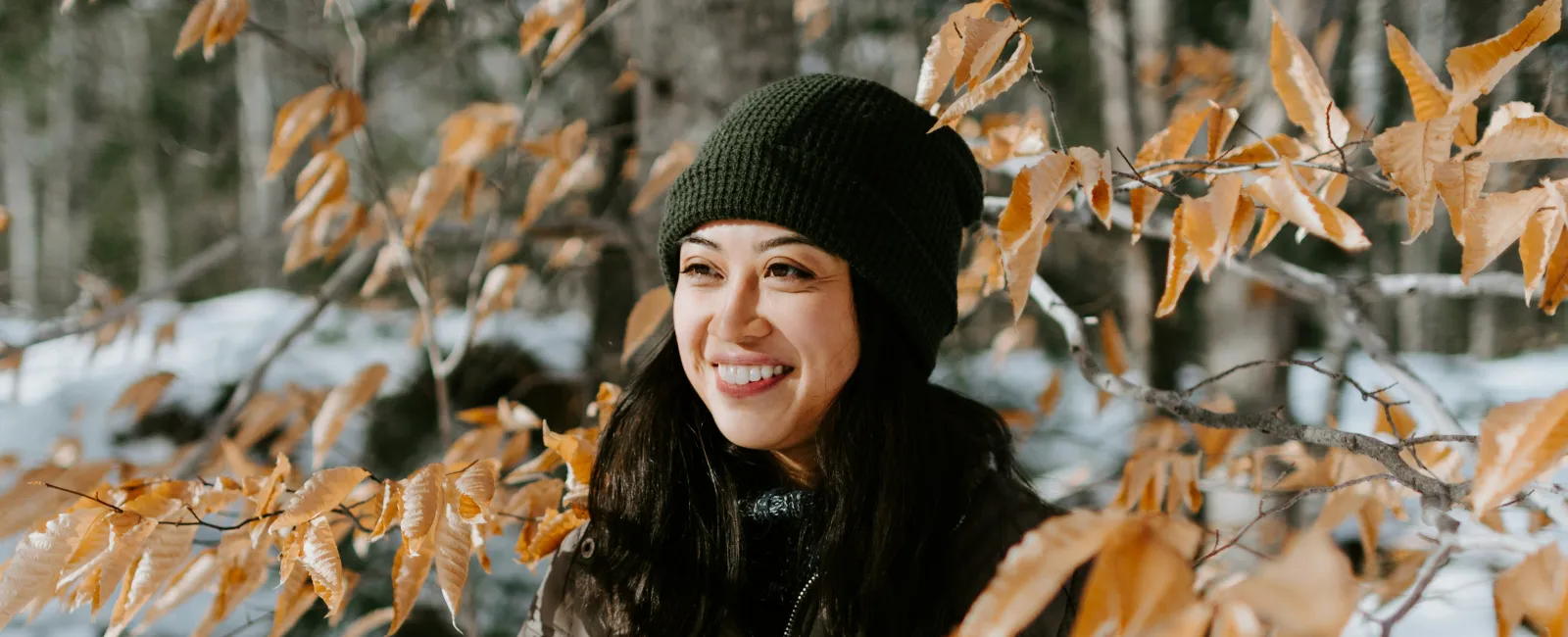 Smiling woman in winter jacket and green beanie standing among orange autumn leaves with snowy forest background.