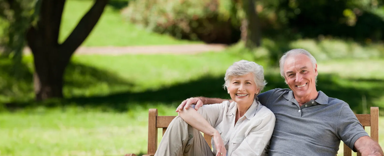 Smiling senior couple sitting on a bench in a park, enjoying a sunny day surrounded by greenery.