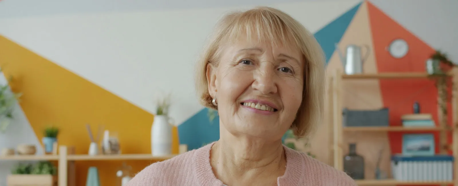 Smiling elderly woman in pink sweater posing indoors with colorful geometric wall and shelves in background