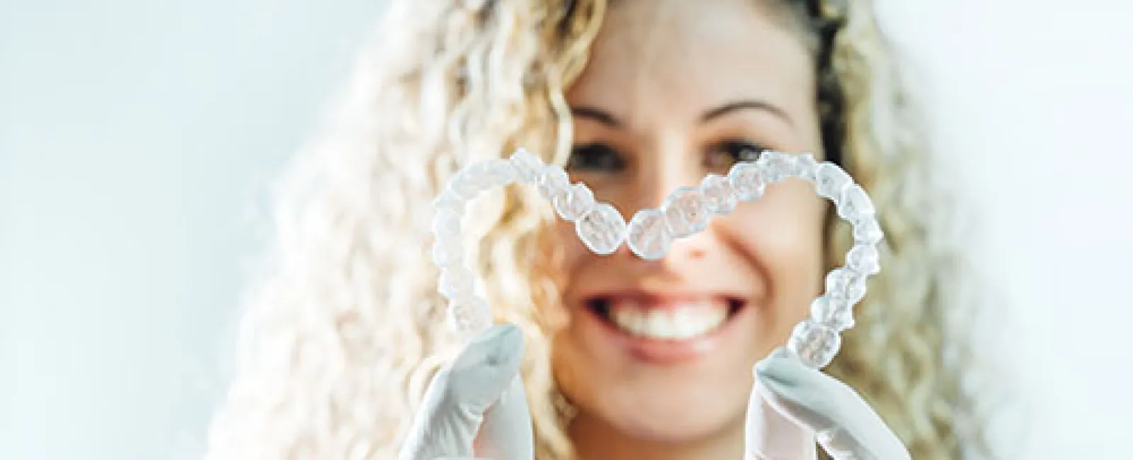 Smiling woman with curly hair holds clear dental aligners shaped like a heart wearing white gloves.