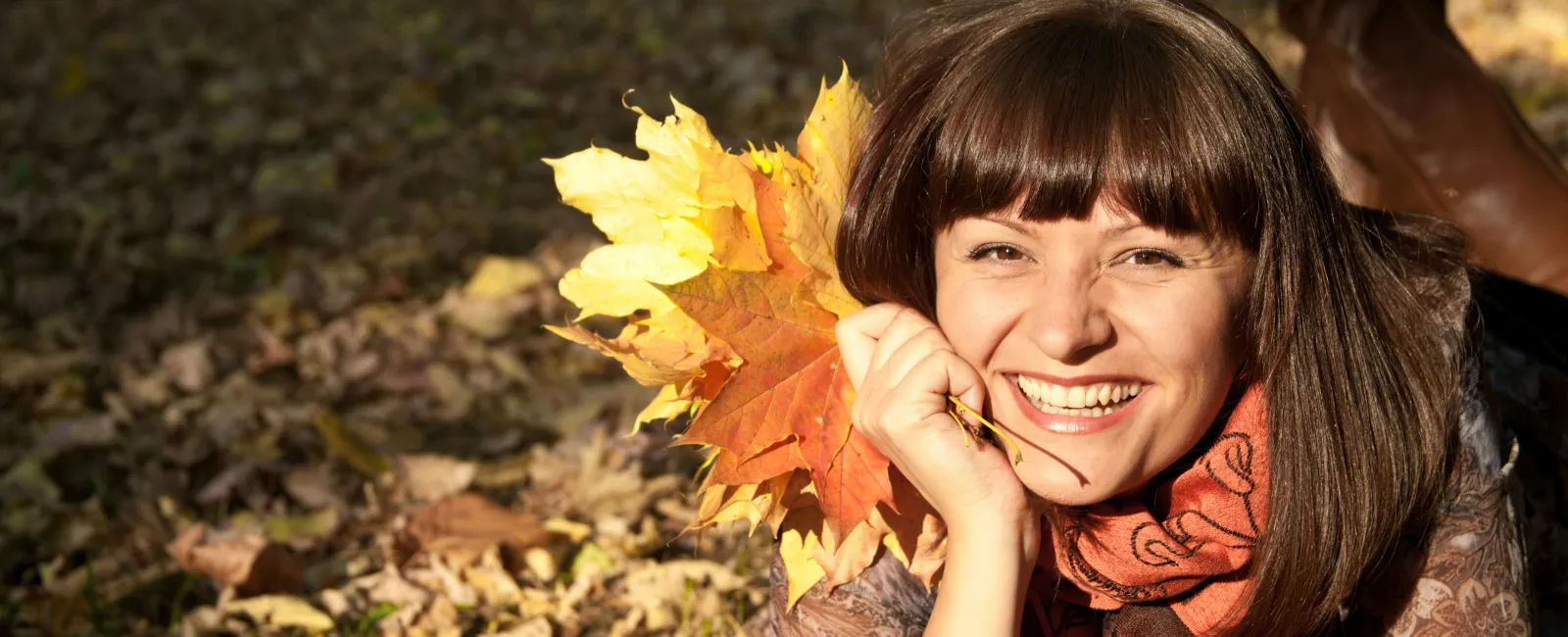 Smiling young woman lying on autumn leaves holding colorful fall foliage in a park on a sunny day