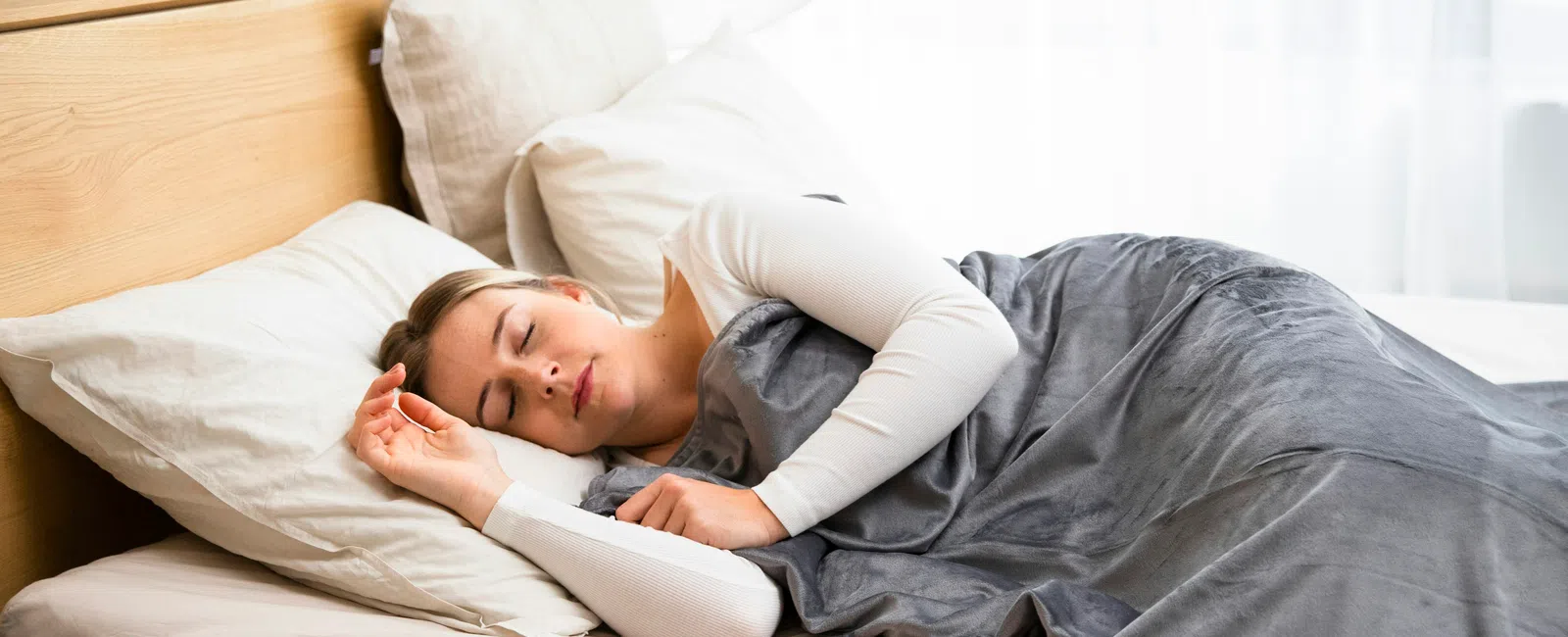 Woman peacefully sleeping in bed covered with a grey blanket and white pillows in a bright room.