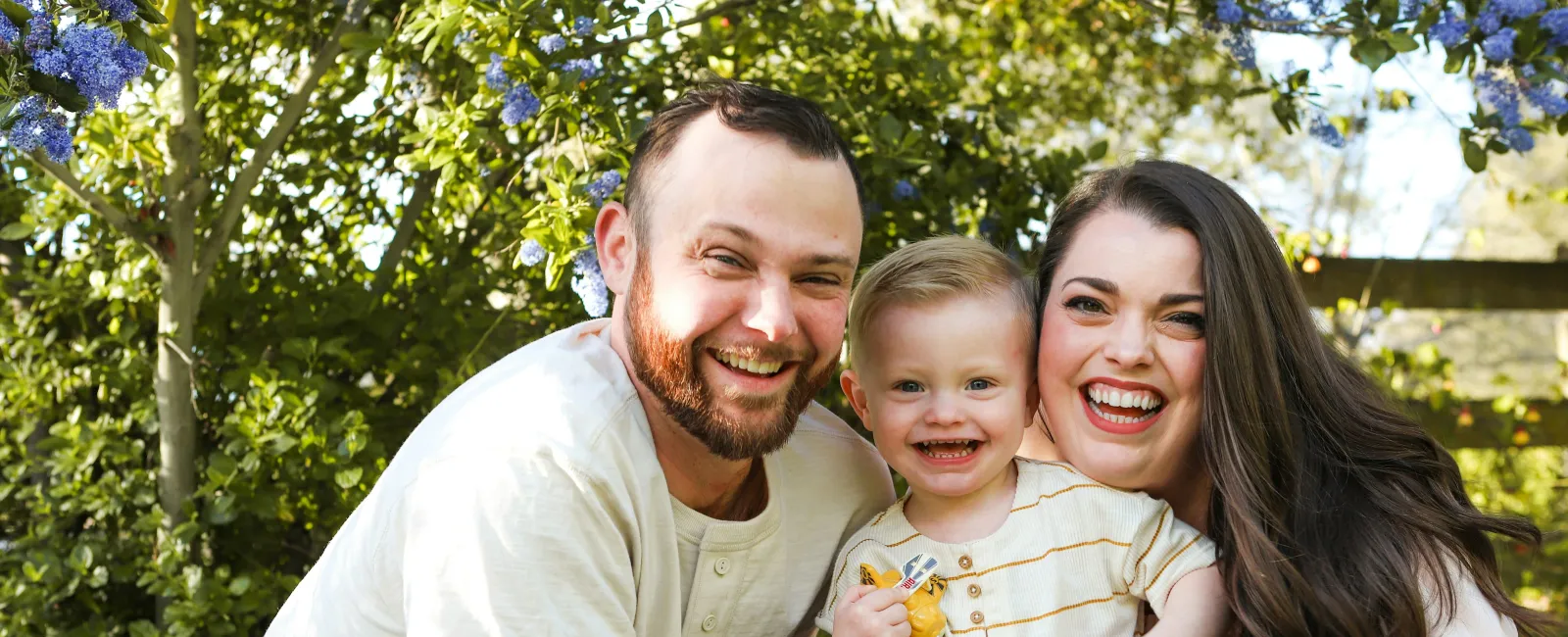 Smiling young family of three outdoors with green foliage and purple flowers in background on sunny day