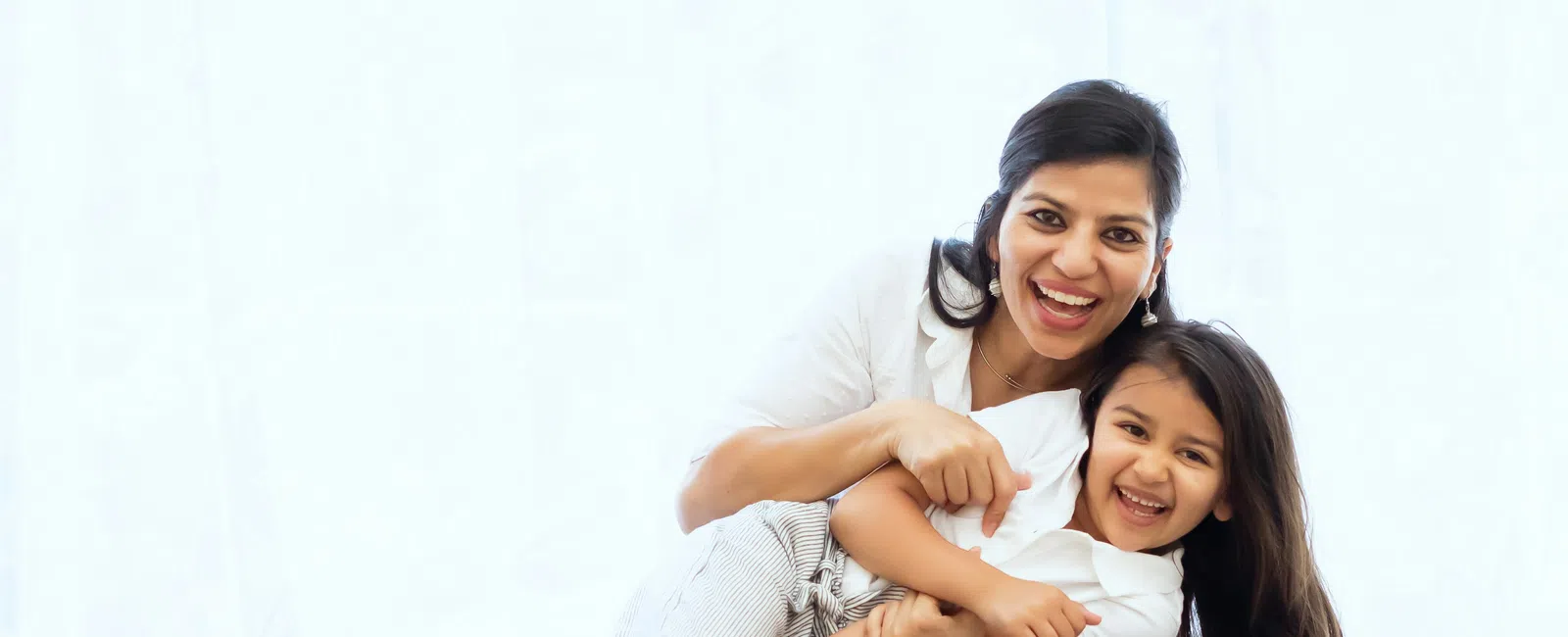 Smiling mother hugging laughing daughter sitting on gray couch with bright white background