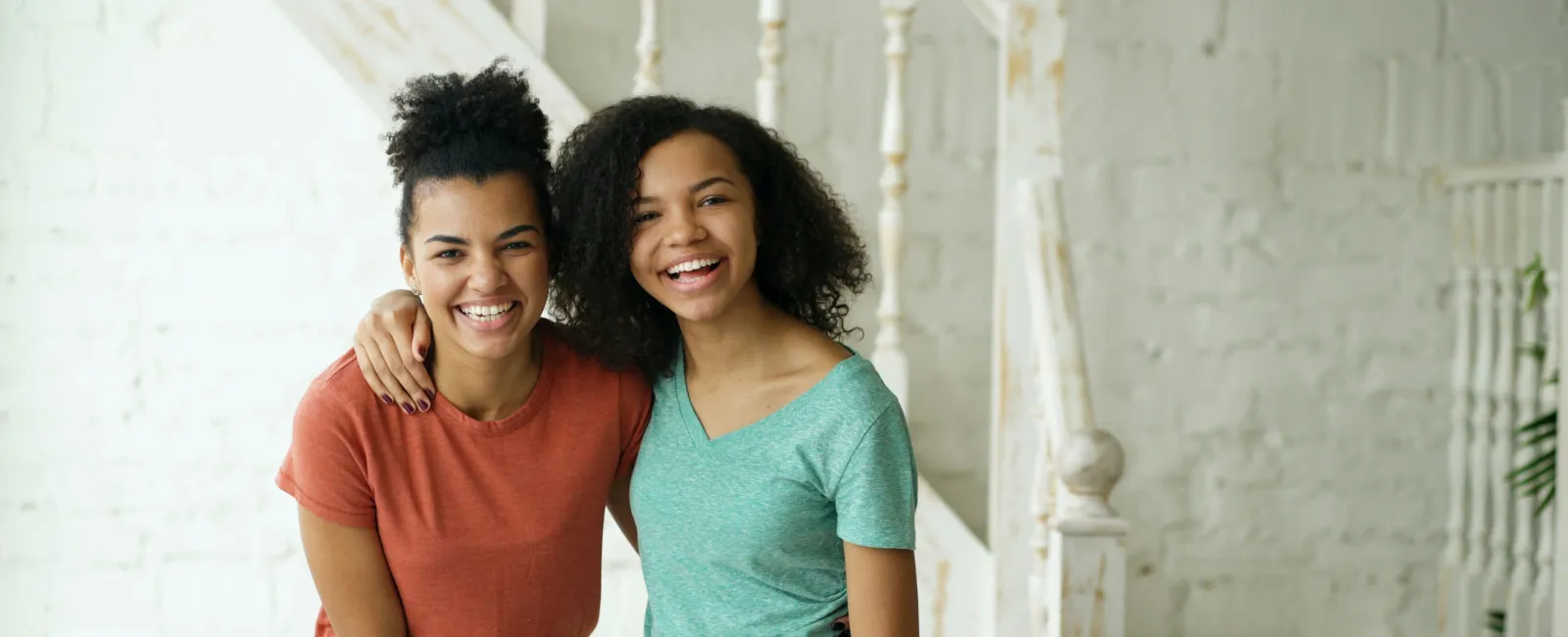 Two cheerful young women in casual clothes smiling and embracing indoors near white stairs and walls.