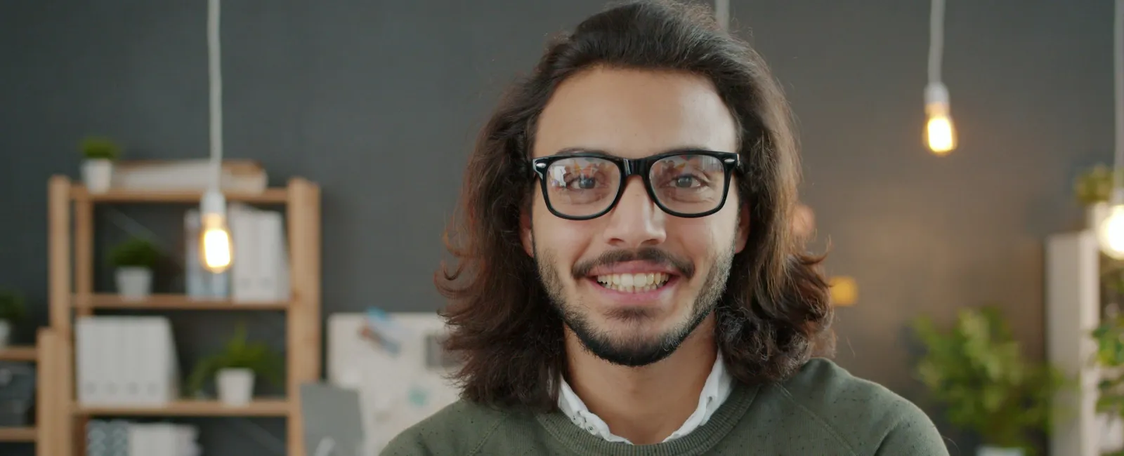 Smiling man with glasses and long hair in a cozy, modern office with shelves and hanging light bulbs.