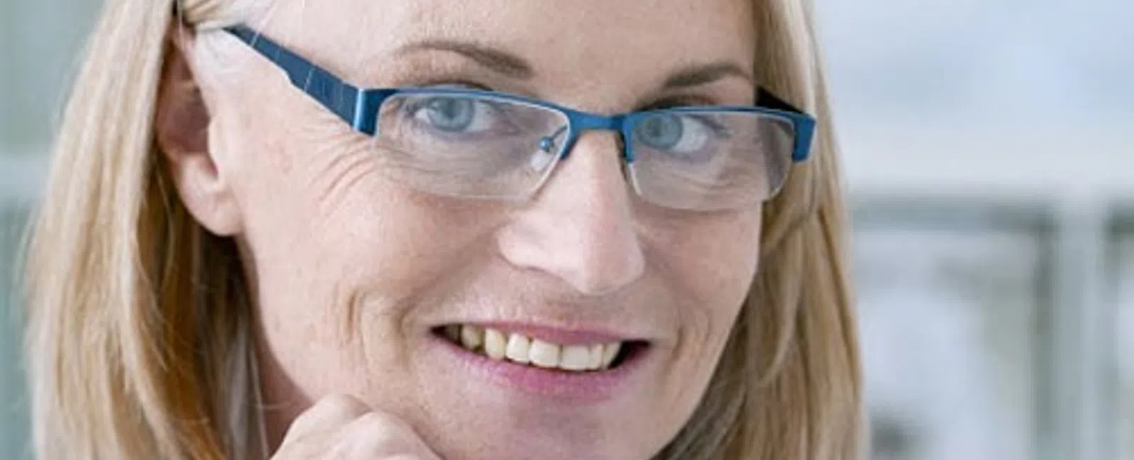 Smiling middle-aged woman with glasses and blonde hair resting her chin on her hand against a blurred background