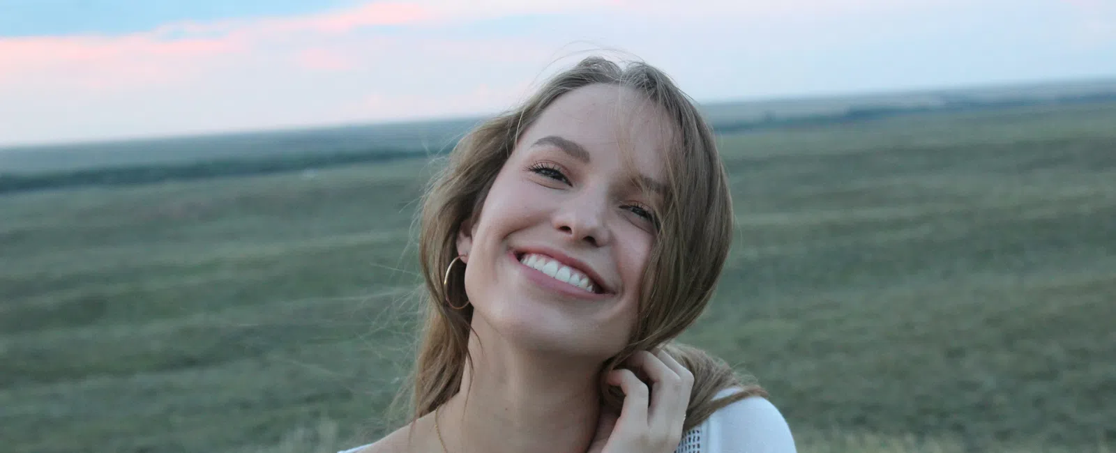 Smiling woman in white lace top standing in open field during sunset with soft pink sky.