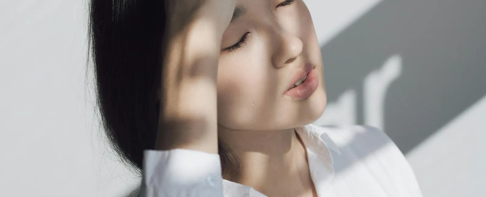 Young woman in white shirt resting her head with closed eyes in soft natural light and shadow.