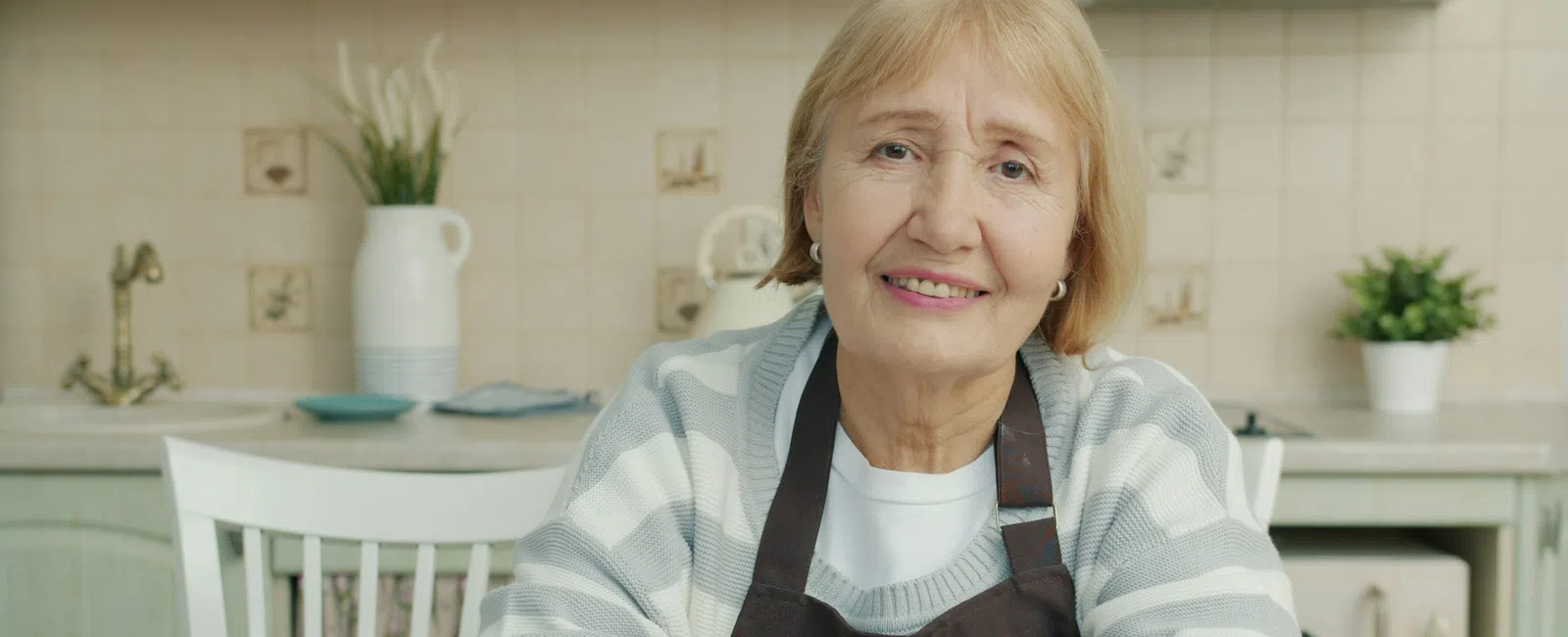 Smiling elderly woman in striped sweater and apron sitting at kitchen table with plants and crockery in background