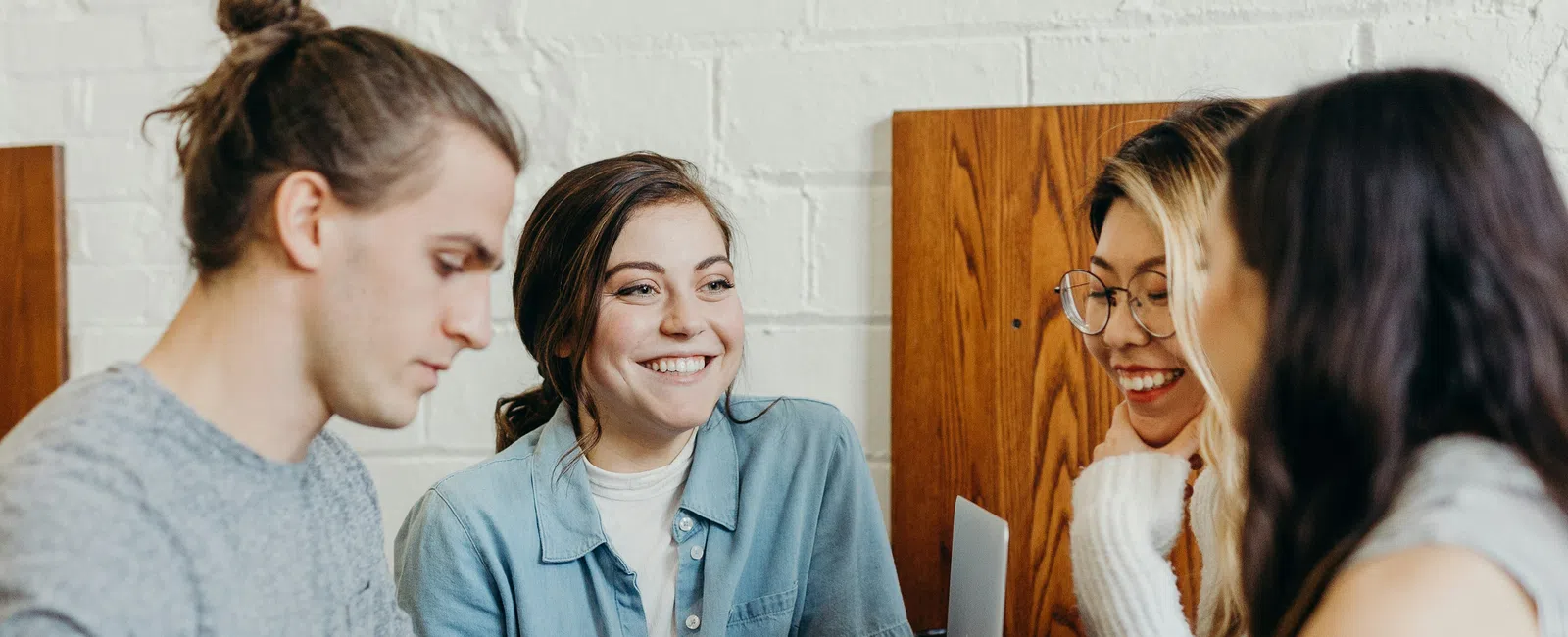 Group of four young adults working and chatting together at a wooden table in a casual cafe setting.