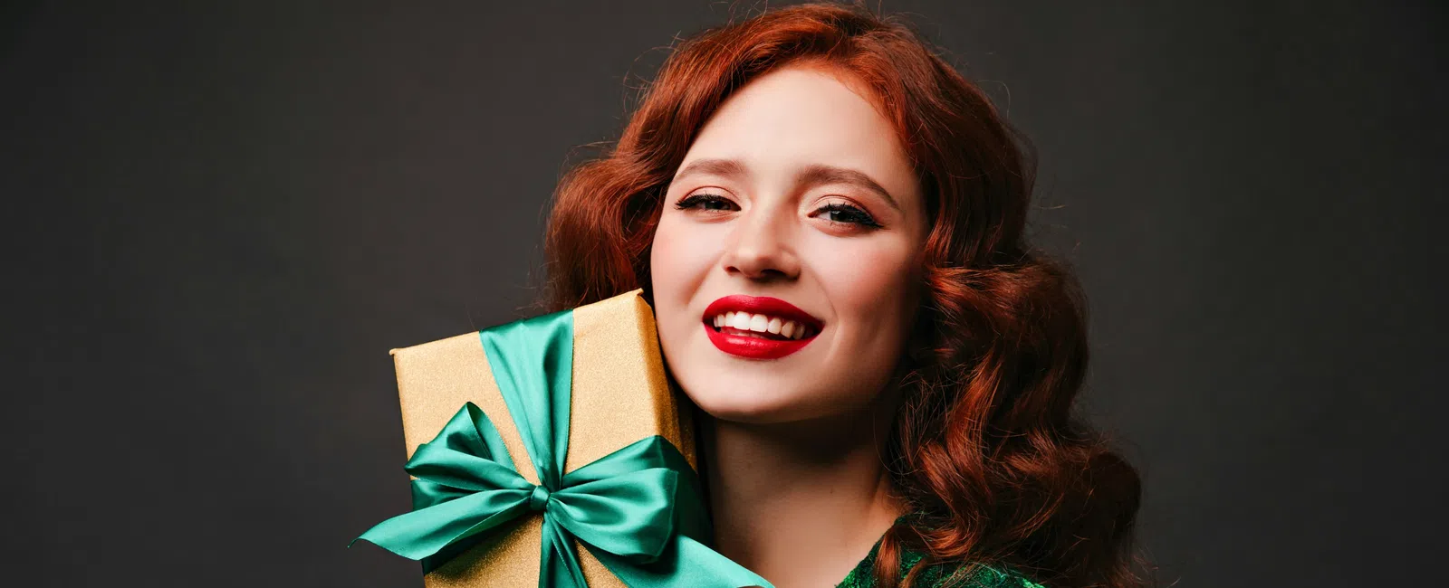 Smiling red-haired woman in green dress holds a gold gift box with a teal ribbon against a dark background.